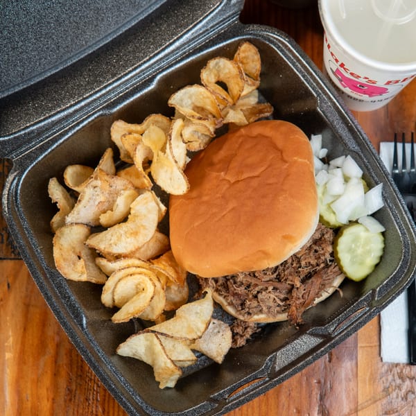 Blake's Barbecue pulled pork sandwich with curly fries, pickles, and onions in a to-go container.