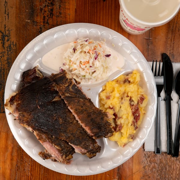 Blake's Barbecue ribs, coleslaw, and mashed potatoes on a white plate with a drink and cutlery.