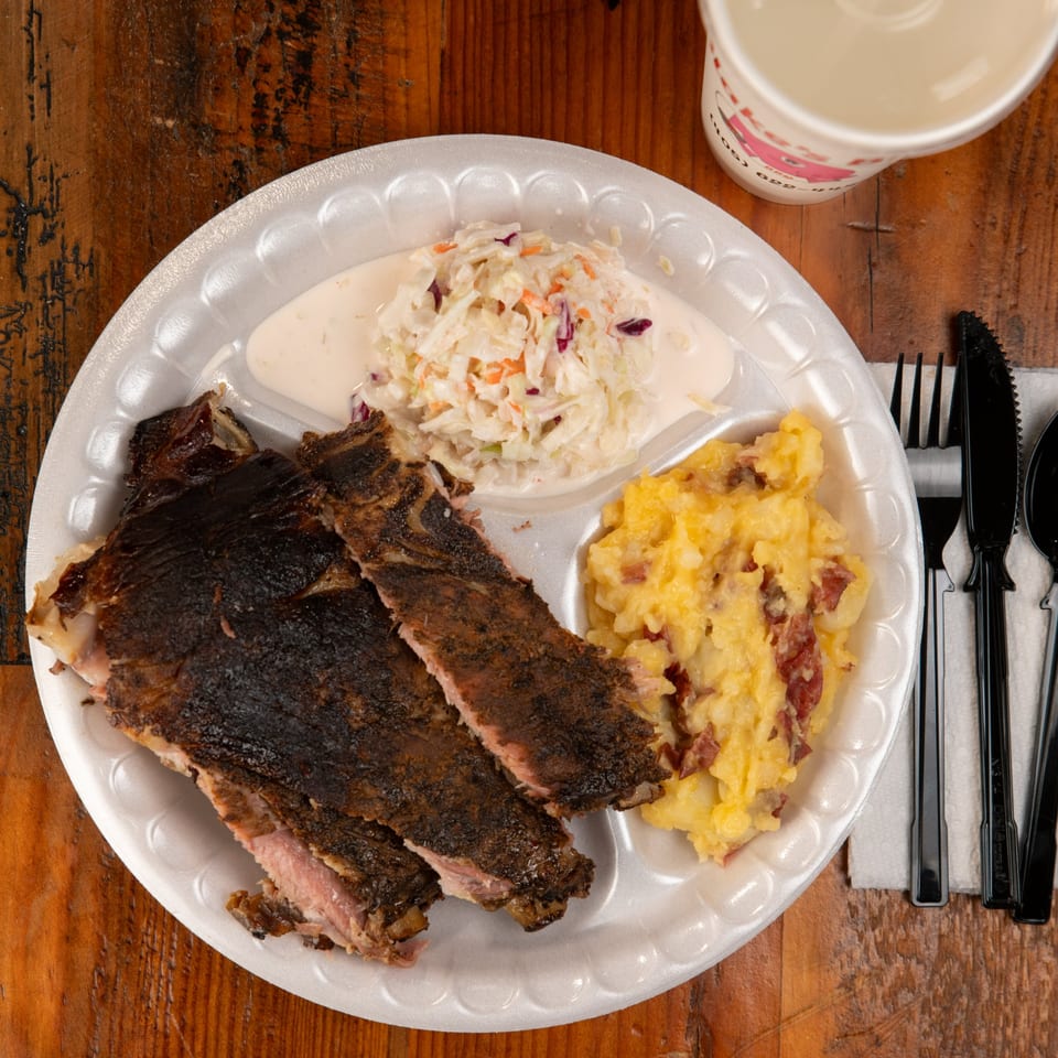 Blake's Barbecue ribs, coleslaw, and mashed potatoes on a white plate with a drink and cutlery.