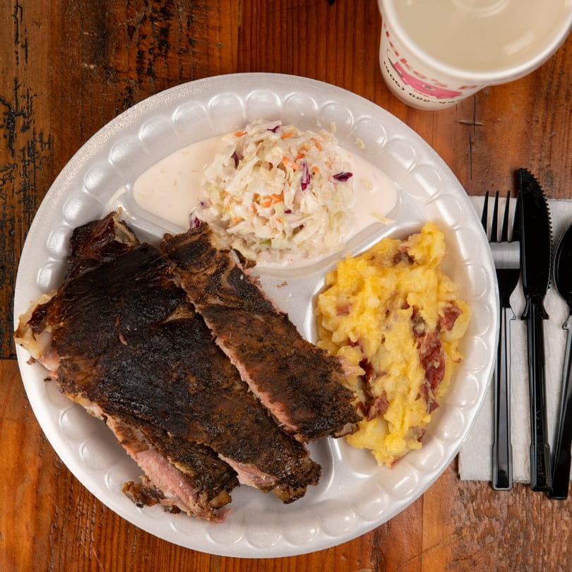 Blake's Barbecue ribs, coleslaw, and mashed potatoes on a white plate with a drink and cutlery.