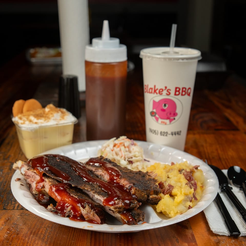Blake's Barbecue ribs, coleslaw, and potato salad on a plate with a drink and banana pudding.